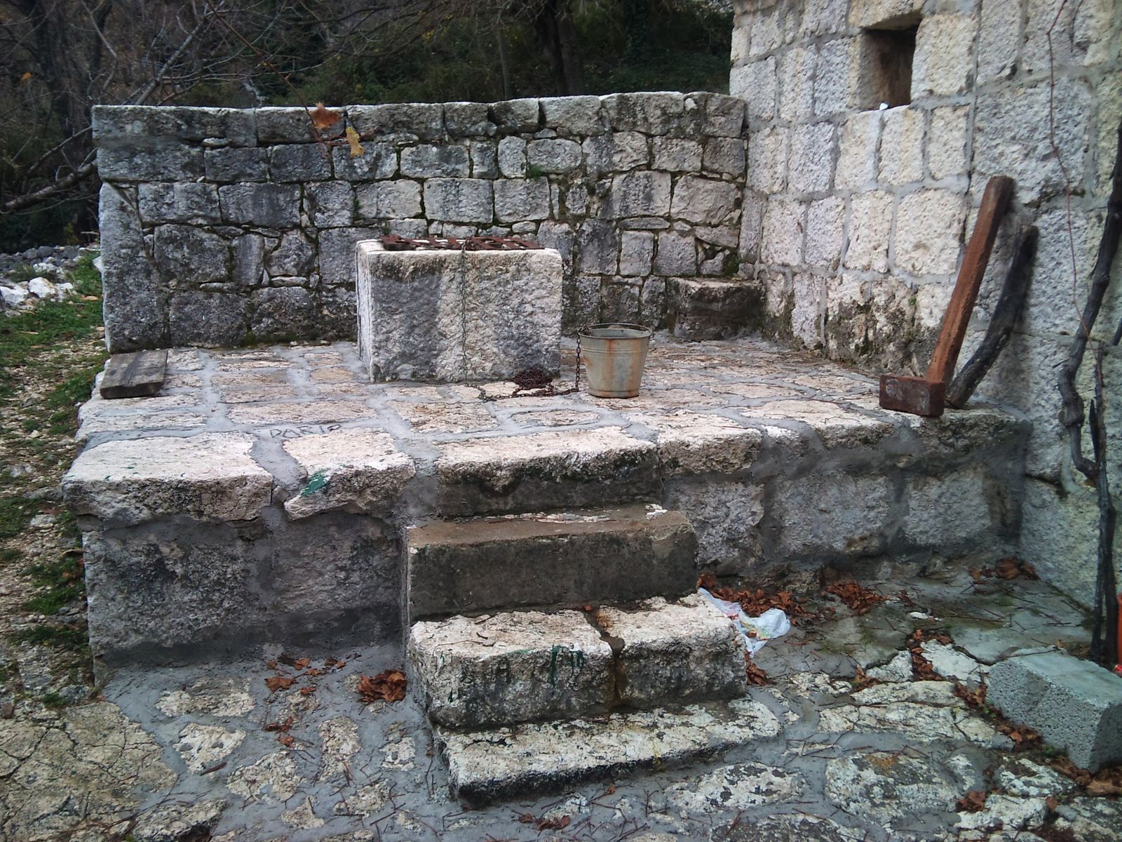 Old house cistern next to a stone house: a raised stone platform with steps and a square stone well-head with a metal grate, a bucket and a chain